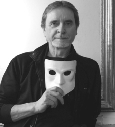 A man holding a white mask sits in front of a mirror, wearing a dark jacket. The image is in black and white.