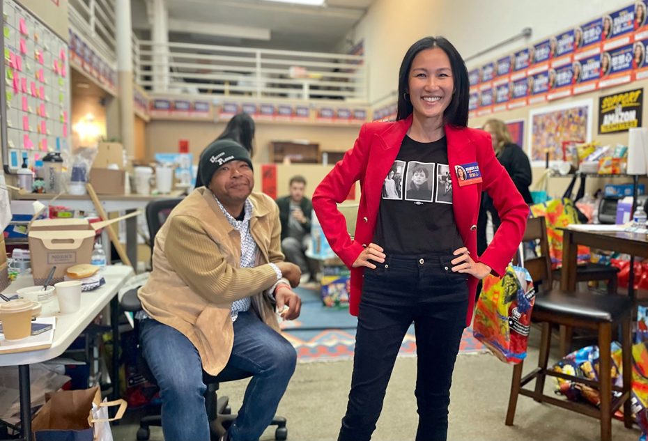 A woman in a red blazer stands smiling in a busy room with campaign posters, snacks, and volunteers. A man is seated at a table nearby.