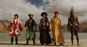 Five people in colorful, theatrical costumes stand on a beach with mountains and a cloudy sky in the background.