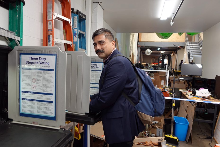 A person with a backpack stands at the voting booth in a cluttered room, ready to play their part in the upcoming election.