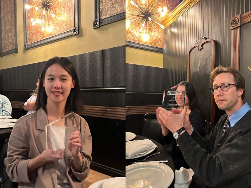 Two people holding triangular glass awards while seated at a dinner table in a restaurant with ornate decorations.