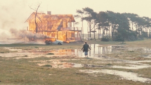 A person walks away from a burning house as flames and smoke rise. Nearby, there is a car close to the fire, and some trees are in the background.