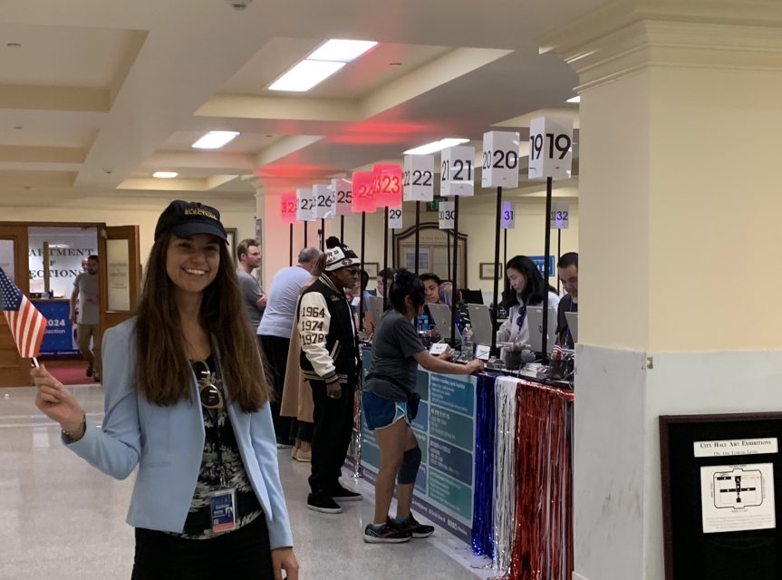 A woman in a blue blazer and cap holds a small flag in a room with people and numbered voting booths.
