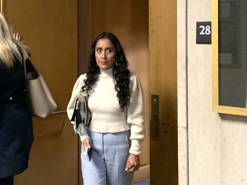 A woman with long curly hair exits a door numbered 28, holding a phone and documents, wearing a white sweater and light blue pants.