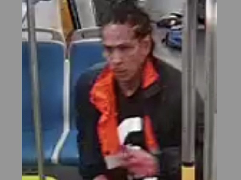 Person seated on BART, wearing a black jacket with an orange interior and a dark shirt. Several empty blue seats are visible around them.
