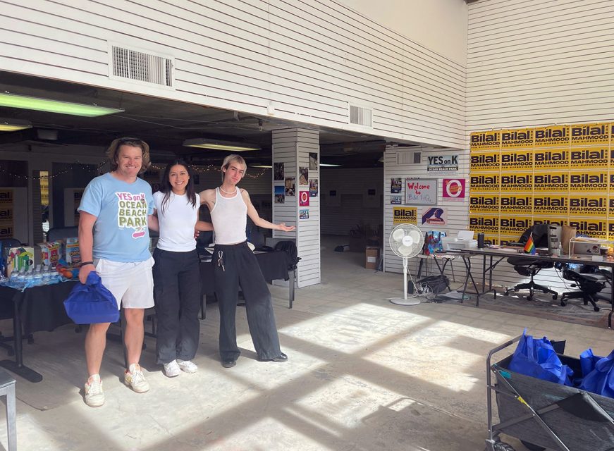 Three people stand in a room with campaign posters on the wall and tables with supplies. They pose and smile for the photo.