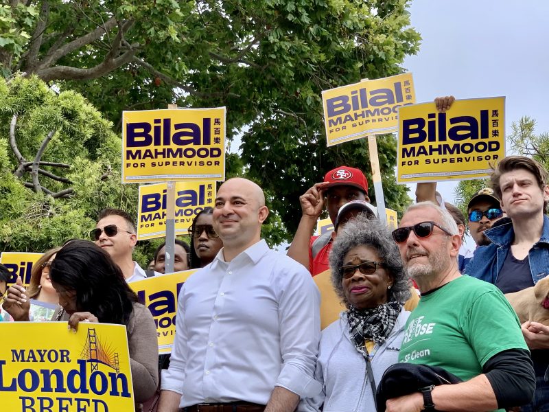A group of people stand together holding "Bilal Mahmood for Supervisor" signs. Some wear sunglasses; others hold smartphones. Trees are in the background.