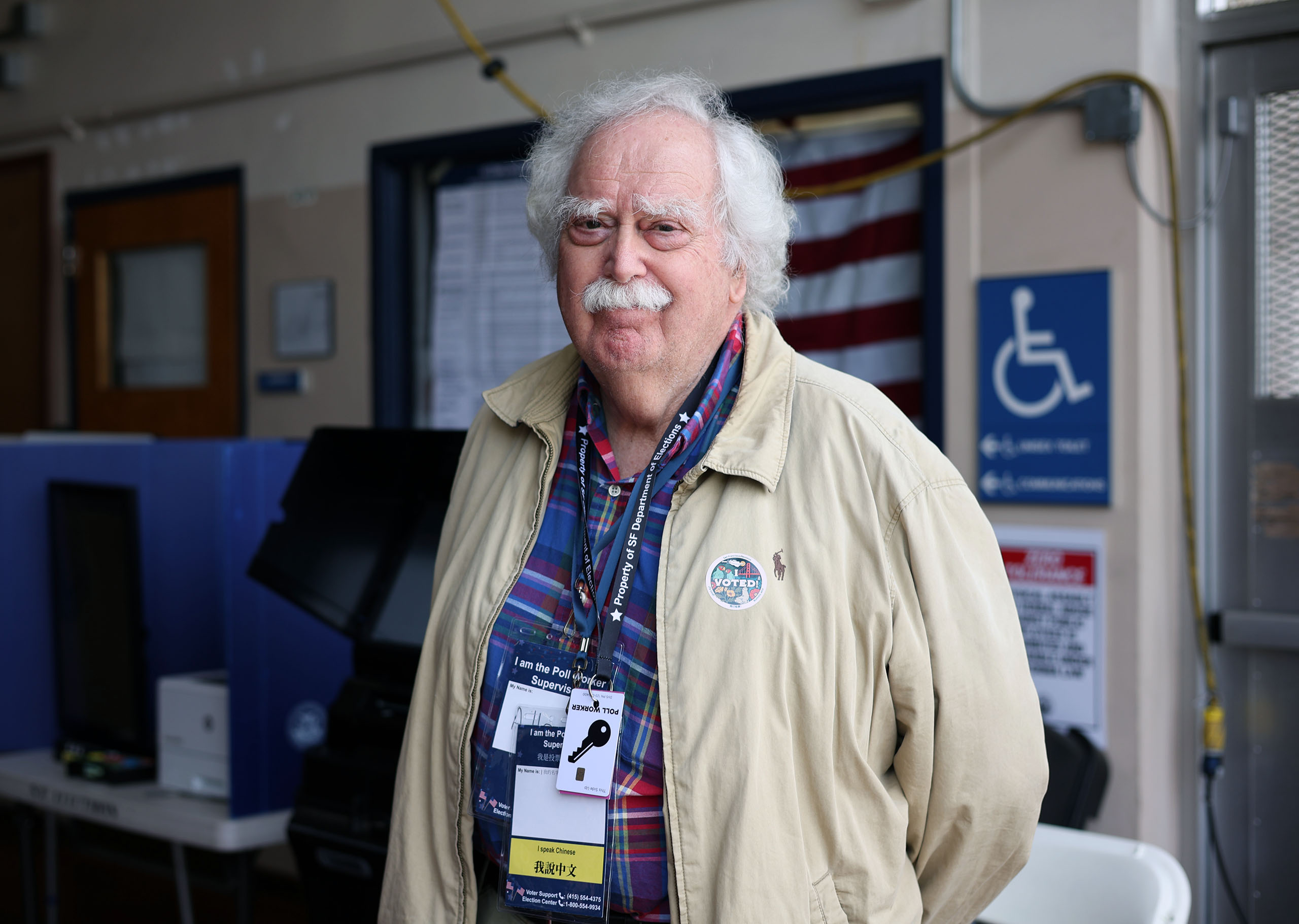 Elderly man with a white mustache wearing a plaid shirt, beige jacket, and ID badge, standing in a room with a blurred American flag and various equipment in the background.