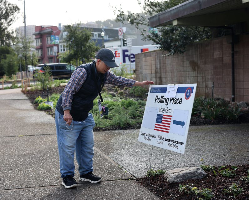A person adjusts a polling place sign outside, surrounded by greenery and buildings.