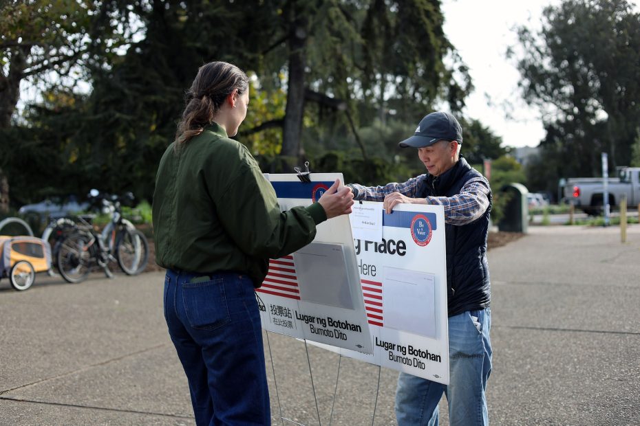 Two people set up a voting sign outdoors on a paved area with bikes and trees in the background.