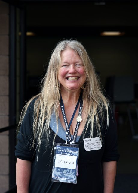 Smiling person with long hair, wearing a black top and name badge, stands in front of a building.