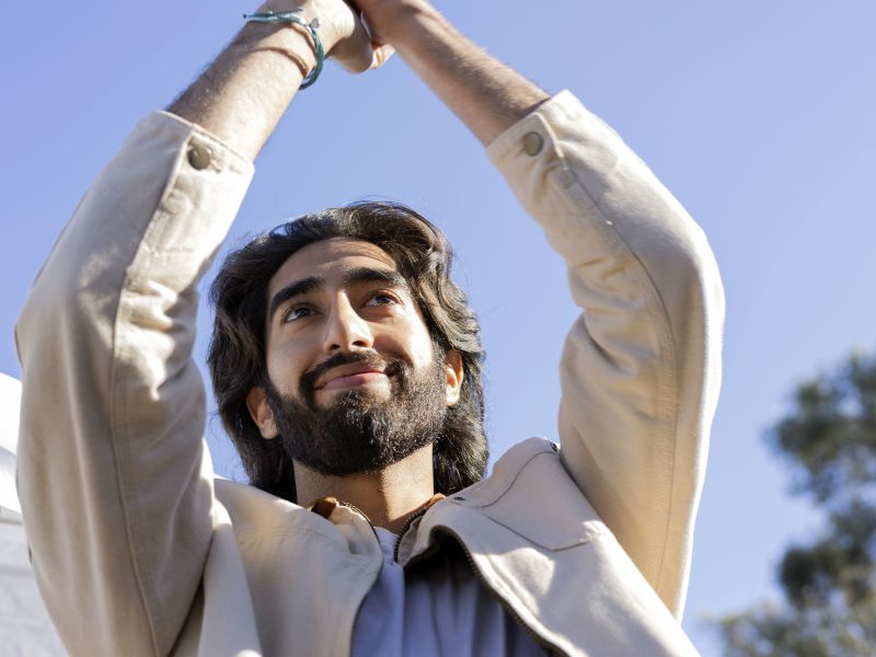 With long hair and a beard reminiscent of Dev Patel, the person raises their arms, smiling against a clear blue sky.