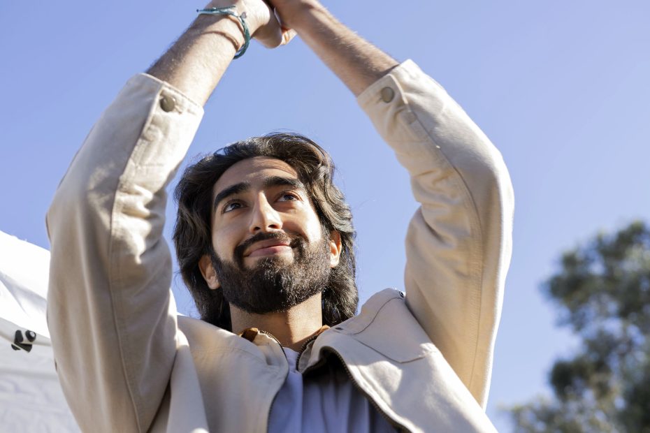 With long hair and a beard reminiscent of Dev Patel, the person raises their arms, smiling against a clear blue sky.