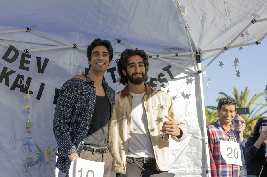 Two people, each wearing numbered tags, stand together under a tent at an outdoor event. Among the participants in the background, Dev Patel can be seen holding a camera.