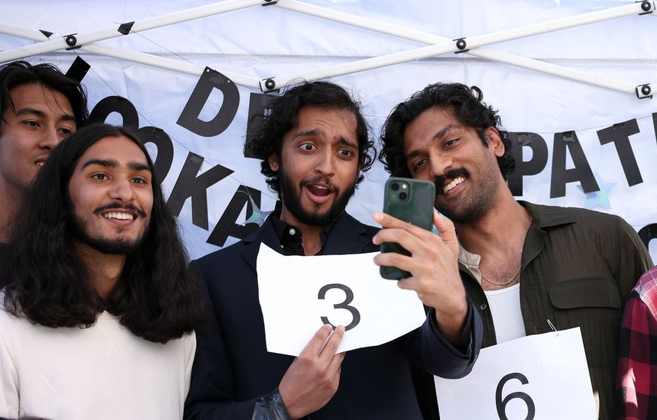 Three individuals pose for a selfie holding numbered signs under a canopy with a partially visible banner.