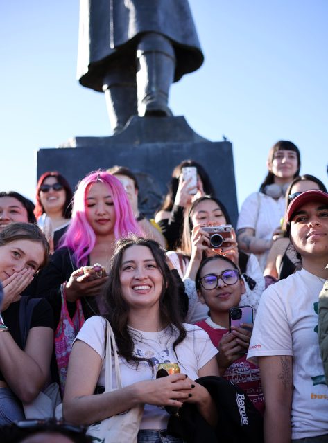 A group of people, some with cameras and phones, gather in front of a large statue under a clear blue sky, hoping for a glimpse of Dev Patel rumored to be filming nearby.
