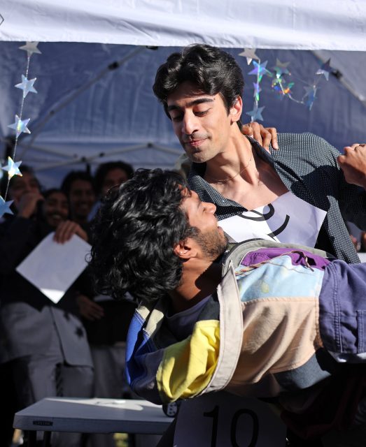 Two men engage in a playful dance under a white tent, with one dipping the other. Star decorations hang above, and people are visible in the background.