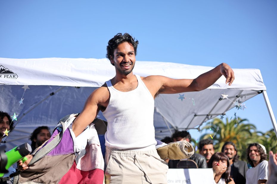A person in a white tank top holds open a colorful coat, smiling. They are standing in front of a white tent, with a crowd of people and trees in the background.