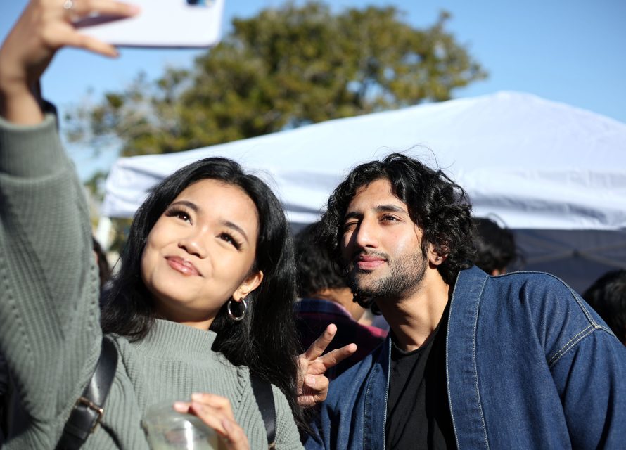 Two people take a selfie outdoors. The woman holds a phone while the man gestures a peace sign. A tent and trees are visible in the background.