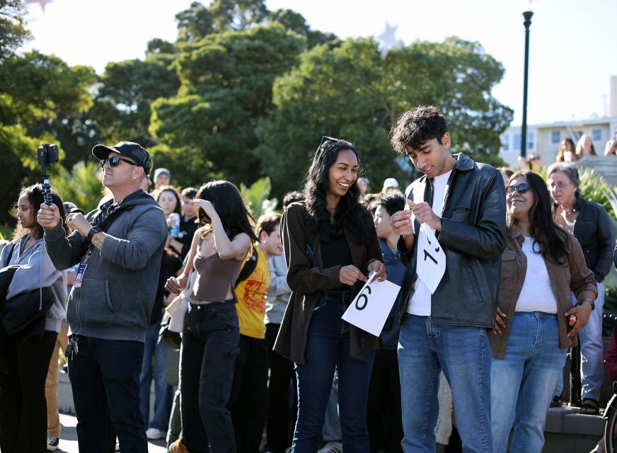 People in an outdoor setting, some holding numbered signs, appear engaged in an event. Trees and buildings are visible in the background.