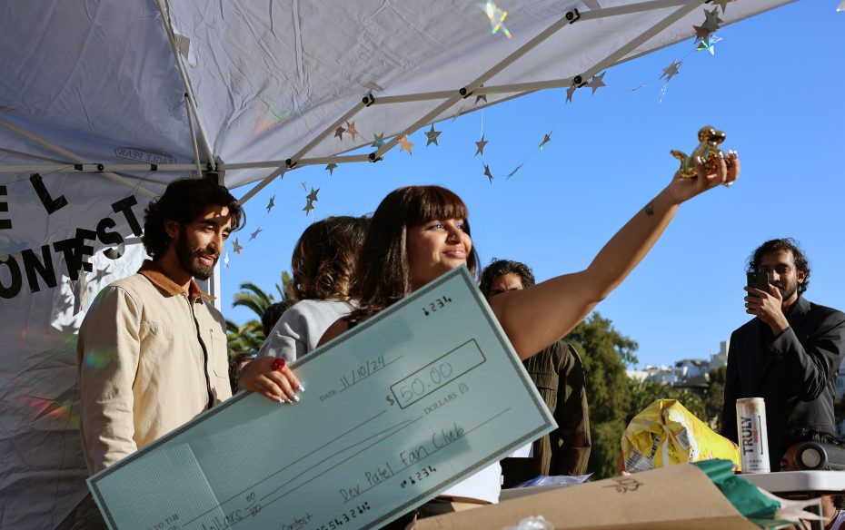 A woman holds up a trophy while others look on. She is also holding a large check. They are outdoors under a decorated canopy.