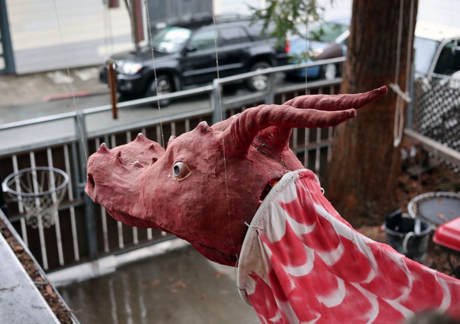 A red dragon head with horns and a scaled body resembling fabric is displayed outdoors, suspended on strings in a residential area.