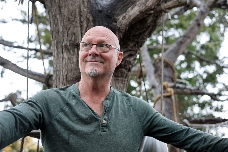 A man with glasses and a green shirt smiles while sitting on a swing in front of a large tree.