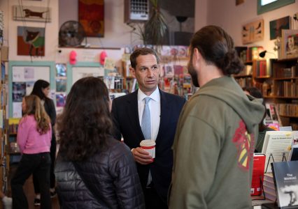 A man in a suit holds a drink and talks to two people in a bookstore. Shelves and patrons are visible in the background.
