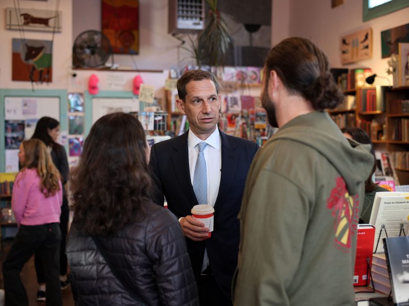 A man in a suit holds a drink and talks to two people in a bookstore. Shelves and patrons are visible in the background.