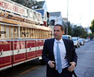A man in a suit walks briskly in front of a parked fire truck on a city street.