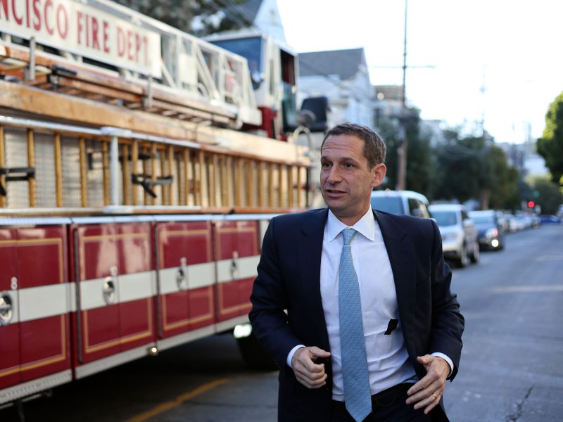 A man in a suit walks briskly in front of a parked fire truck on a city street.