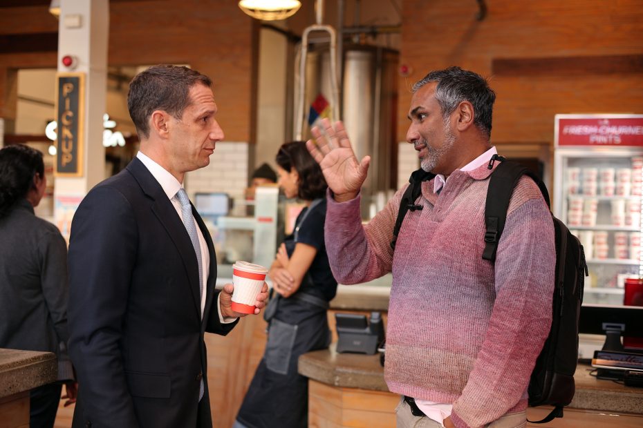 Two people are having a conversation in a cafe. One holds a coffee cup, and the other gestures with a raised hand. Others are in the background near a counter.