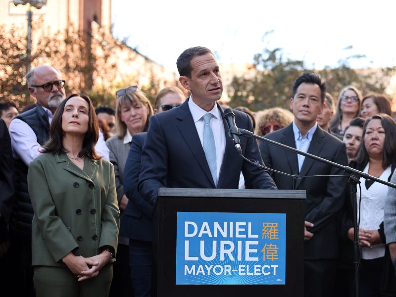A man speaks at a podium labeled "Daniel Lurie Mayor-Elect," surrounded by a group of people outdoors.