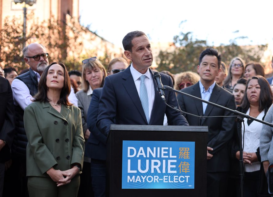 A man speaks at a podium labeled "Daniel Lurie Mayor-Elect," surrounded by a group of people outdoors.