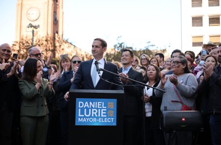 A man stands at a podium labeled "Daniel Lurie Mayor-Elect," surrounded by applauding people. A clock tower is in the background.