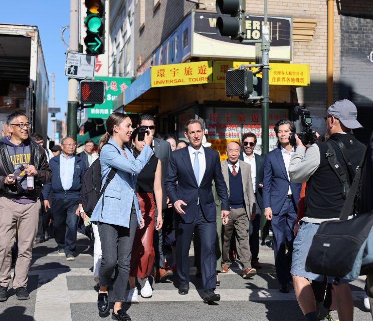 A group of people in business attire walk in an urban area with cameras around. Bright sunlight and buildings with signs in the background.