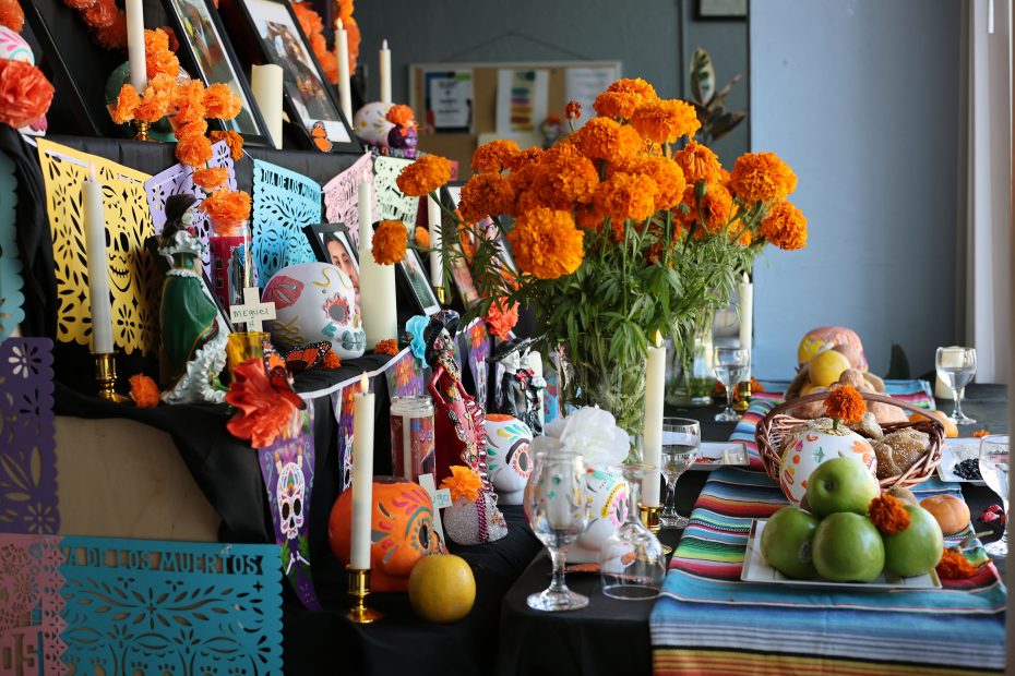 A Día de los Muertos altar with marigolds, candles, decorated skulls, and fruit. Colorful papel picado banners and photos are displayed.