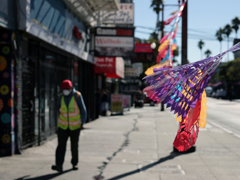 Colorful papel picado decorations flutter above a city street. A person in a reflective vest and hat walks along the sidewalk, with various shop signs visible in the background.