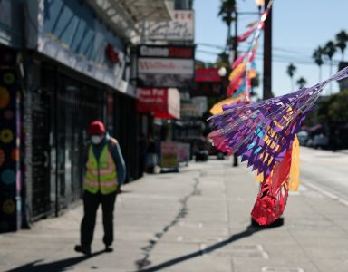 A pedestrian wearing a safety vest and mask walks on a sidewalk lined with colorful papel picado banners, on a sunny day.