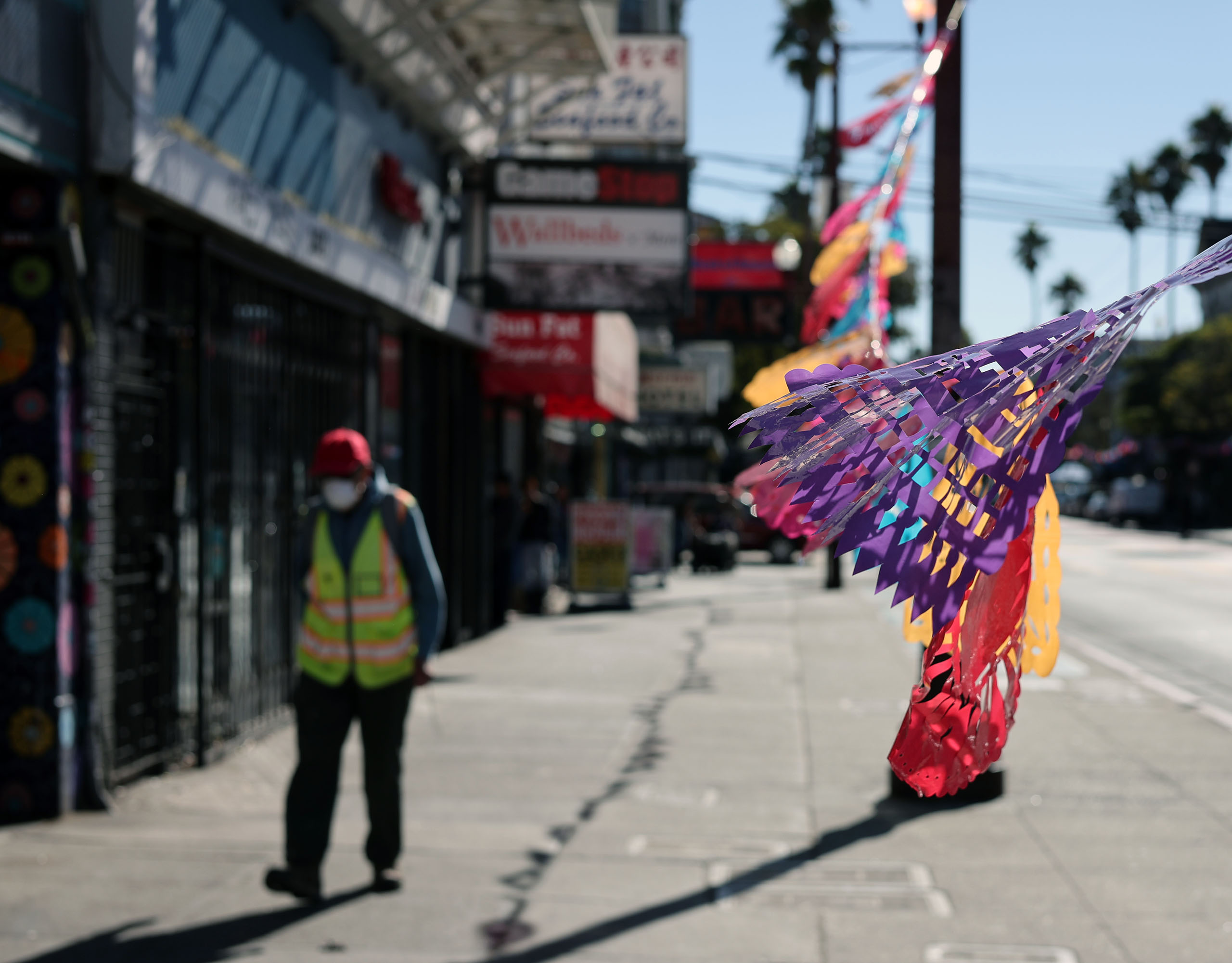A pedestrian wearing a safety vest and mask walks on a sidewalk lined with colorful papel picado banners, on a sunny day.
