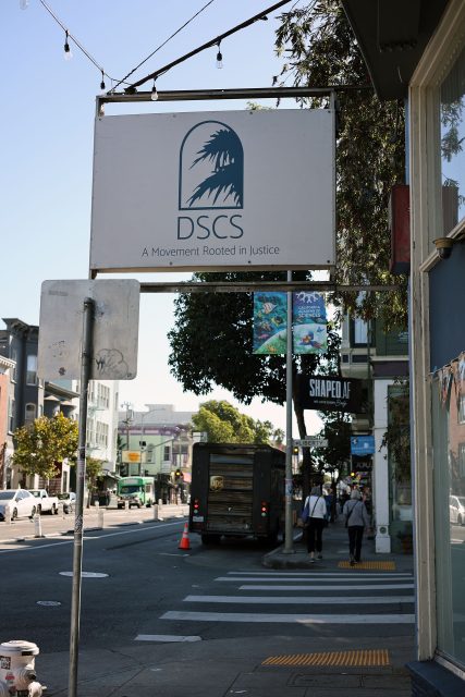 Street view with a sign reading "DSCS A Movement Rooted in Justice" hanging above the sidewalk. Buildings, parked cars, and pedestrians in the background.