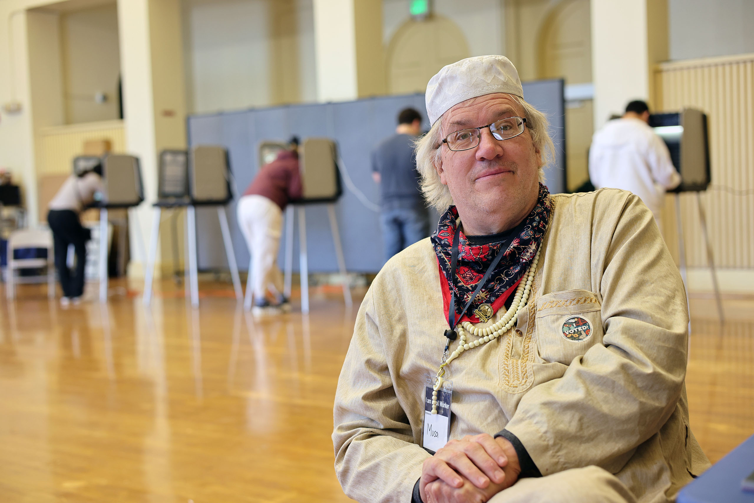A person wearing a hat and glasses sits in a room with people voting at booths in the background.