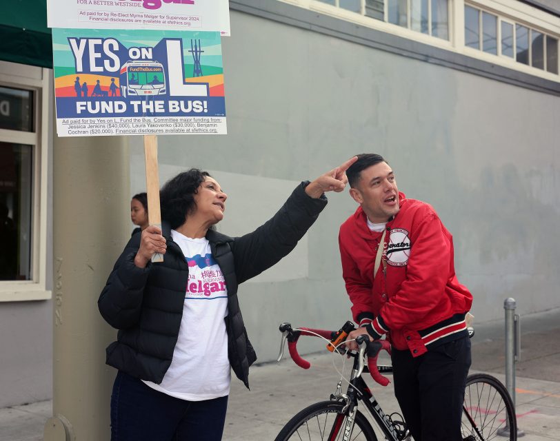 Two people stand near a building; one holds a "Yes on Fund the Bus!" sign, while the other leans on a bicycle.