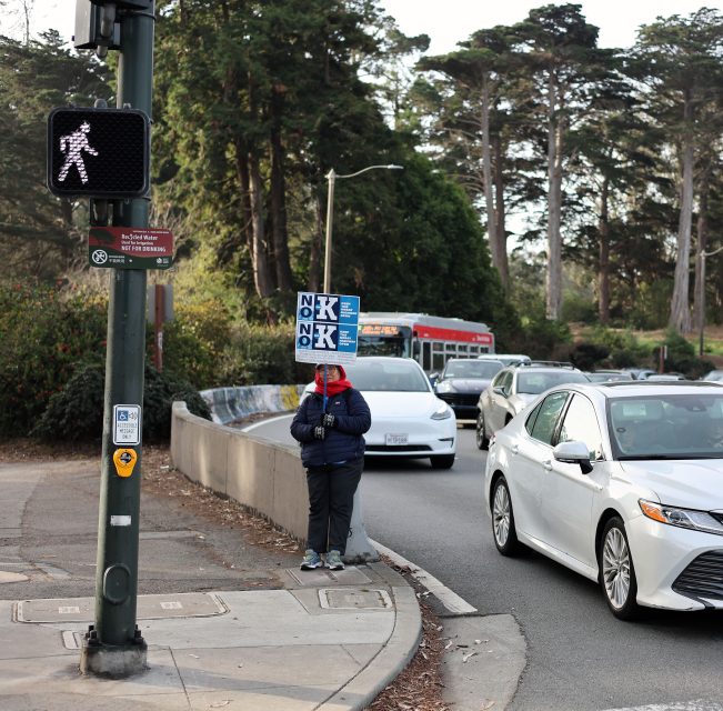 A person stands on a sidewalk holding protest signs with large letters. Cars pass by on the road, and trees are visible in the background.