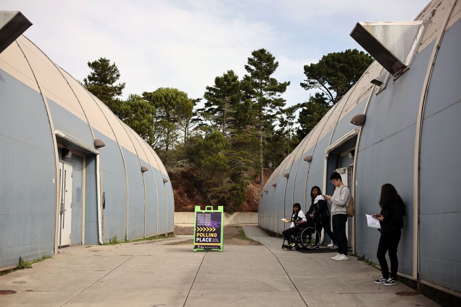People standing outside a polling place, with a sign in the center, flanked by two curved buildings.