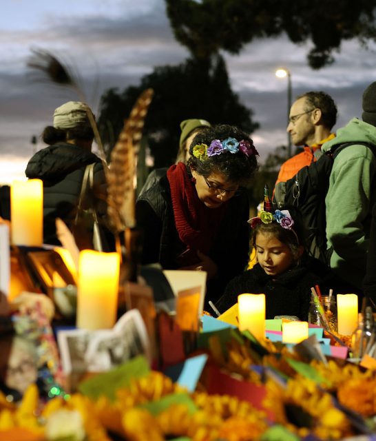 A group of people gather around a table with lit candles and decorative items. A woman and a child focus on something in front of them. The setting appears to be outdoors in the evening.