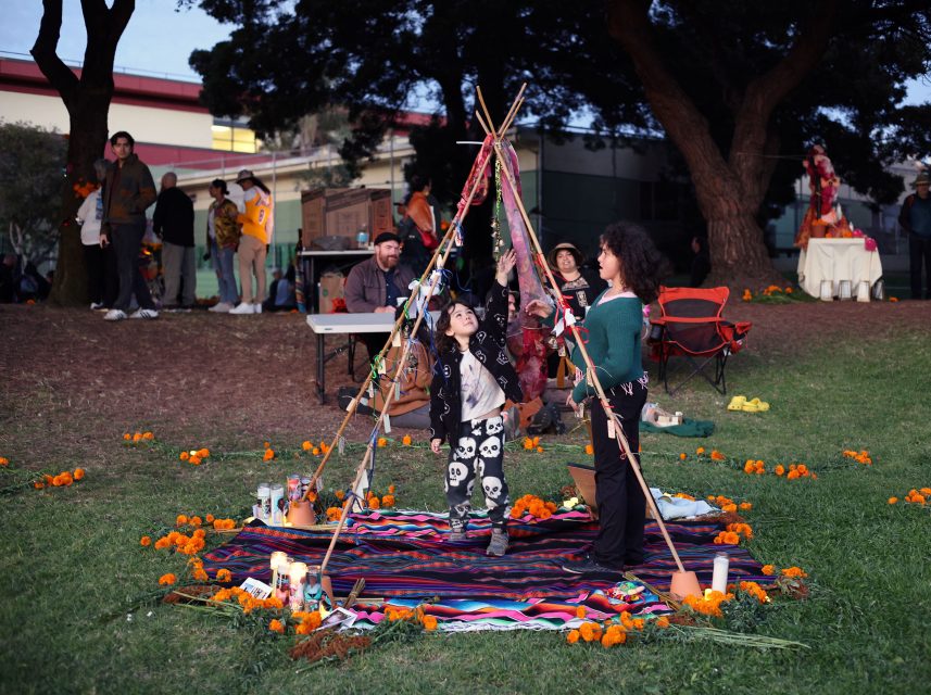 Two children stand under a decorated wooden structure on a colorful blanket, surrounded by marigolds and candles, in an outdoor setting with other people and trees in the background.