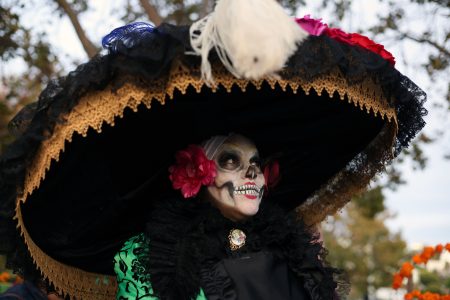 Person dressed as La Catrina with a large hat, face painted in skeleton style, wearing colorful flowers and a decorative outfit at an outdoor event.