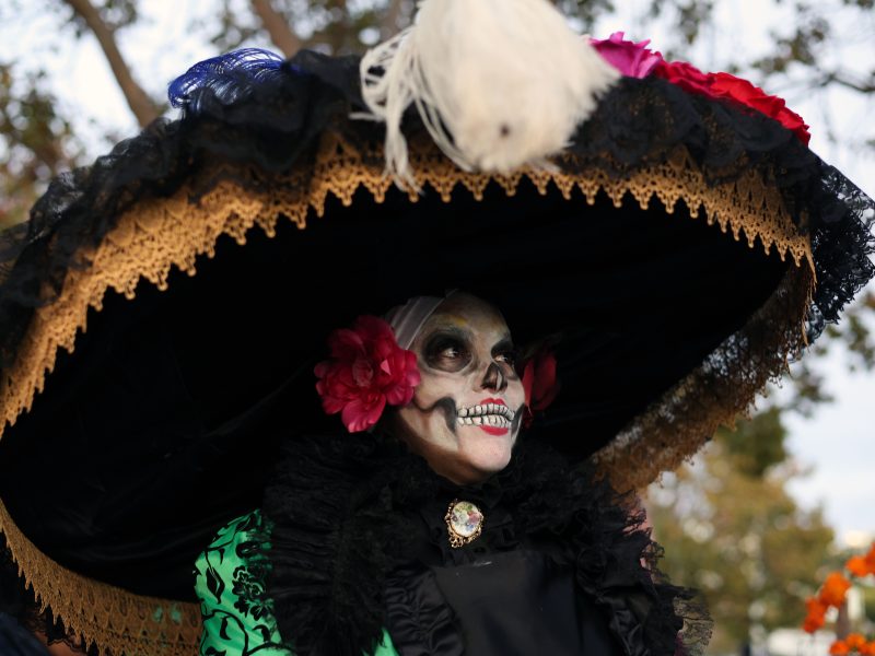 Person dressed as La Catrina with a large hat, face painted in skeleton style, wearing colorful flowers and a decorative outfit at an outdoor event.