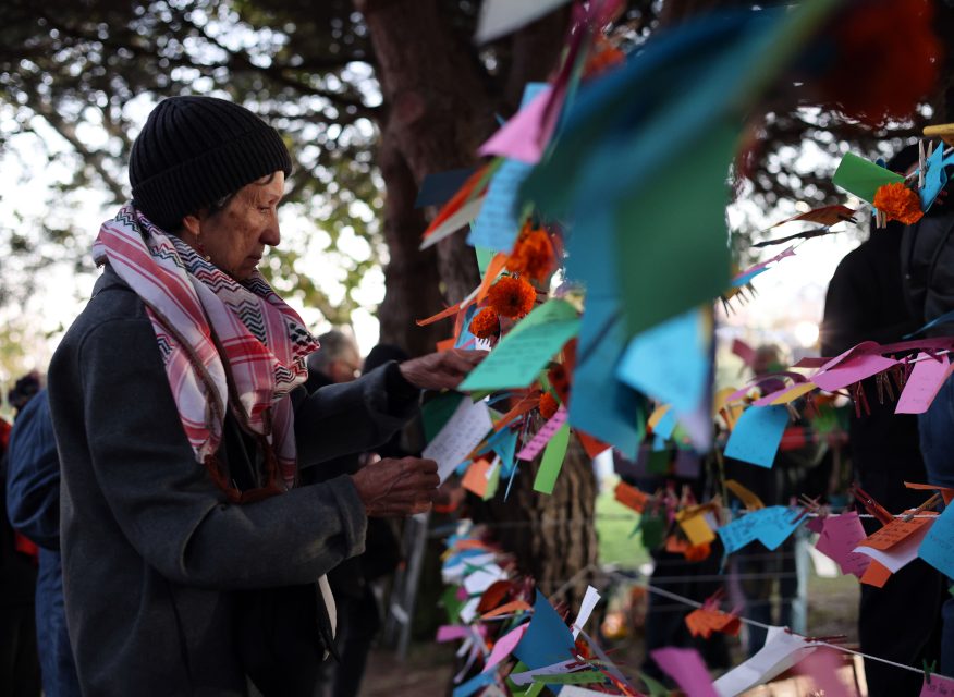 A person in a beanie and scarf reads colorful tags hanging from a tree branch with flowers attached. Día de los Muertos